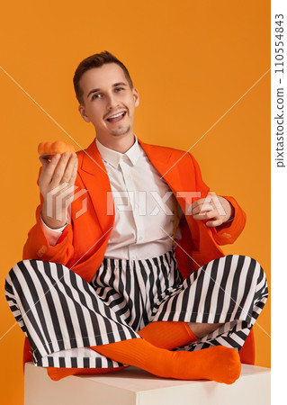 extravagant funny young man in orange jacket and striped pants holding cupcake on orange background. 110554843