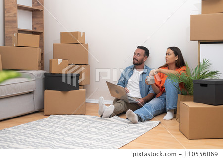 Young people sit on floor with carpet among carton boxes checking availability of decorations for placing in interior of apartment in electronic list on laptop 110556069