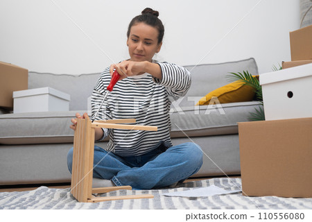 Attractive woman with instrument assembling furniture after shopping at hardware store sitting on floor near soft sofa among collected cardboard boxes of various sizes 110556080