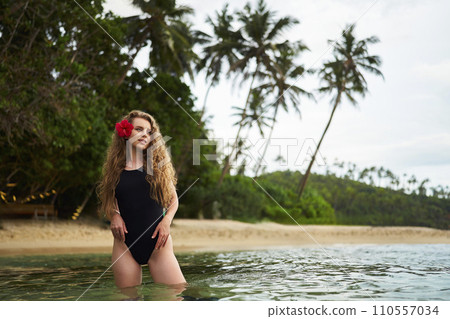 Woman with floral hair accessory wades in tropical sea. Solo traveler enjoys serene beach. Curly-haired female experiences coastal getaway, sunset bathes. Idyllic island leisure, ocean dip moment. Woman with floral hair accessory wades in tropical sea. Solo traveler enjoys serene beach. Curly-haired female experiences coastal getaway, sunset bathes. Idyllic island leisure, ocean dip moment. 110557034
