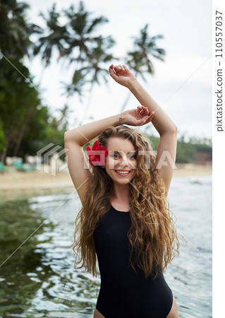 Woman with red hibiscus in hair enjoys tropical sea. Female in black swimsuit at beach escape. Solo traveler explores coast, enjoys summertime swim. Wellness retreat in island paradise. 110557037