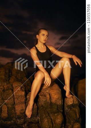 Elegant woman models in black swimsuit on rocky beach at dusk. Athletic female poses, serene seascape background. Swimwear fashion shoot, outdoor lifestyle, summer evening allure. 110557056