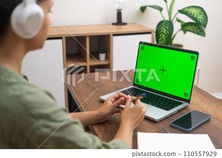Female student in headphones watches online lesson on laptop with chromakey screen woman holds pen preparing to make notes of important information sitting at home side view 110557929