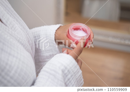 Woman in bathrobe holding open bottle of moisturizing cream near mirror in bathroom lady showing organic cosmetic product for skin pampering in bathroom closeup 110559909