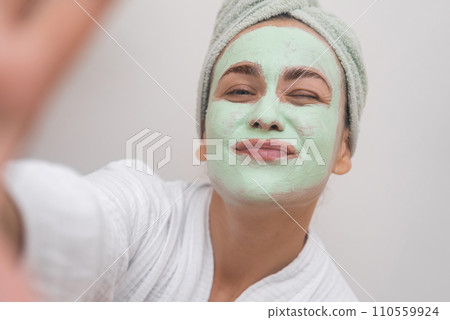Clay Mask Cheer: A bathrobe-clad woman captures her joyful essence in a selfie with a clay mask, beaming at the camera. Clay Mask Cheer: A bathrobe-clad woman captures her joyful essence in a selfie with a clay mask, beaming at the camera. 110559924