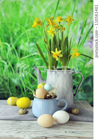 Easter eggs and a bouquet of daffodils in a jug, on a wooden table, against a background of a green lawn, Easter still life 110561401