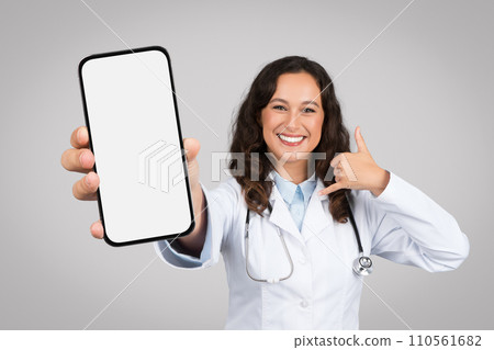 Glad woman doctor in coat showing smartphone with blank screen, call me sign with hand, isolated on grey background Glad woman doctor in coat showing smartphone with blank screen, call me sign with hand, isolated on grey background 110561682