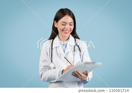 Happy woman doctor writing on clipboard against blue backdrop Happy woman doctor writing on clipboard against blue backdrop 110561724