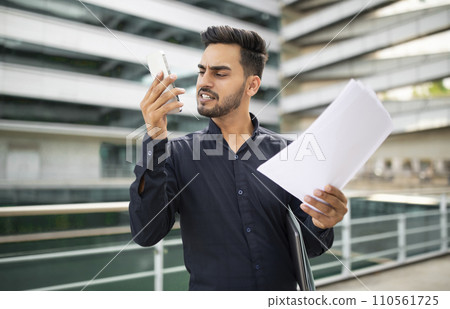 Discontented arabic businessman with business papers talking on phone outdoor Discontented arabic businessman with business papers talking on phone outdoor 110561725