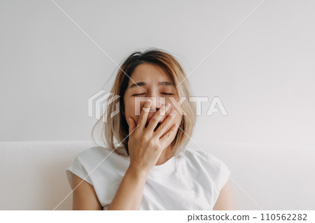 Sleepy face woman yawning and rubbing her eyes in white t-shirt sits on the sofa 110562282