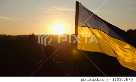 Male soldier stands with raised flag of Ukraine against background of beautiful sunset. Male ukrainian army soldier with a lifted blue-yellow banner in honor of the victory against russian aggression 110562670