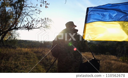 Young male military in uniform walks with waving flag of Ukraine at countryside. Soldier of ukrainian army going with blue-yellow banner outdoor. Concept of victory against russian aggression. 110562678