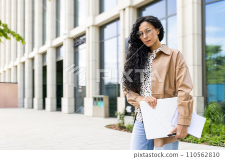 Confident young businesswoman with laptop and notebook standing in front of an office building, exuding professionalism and ambition. Confident young businesswoman with laptop and notebook standing in front of an office building, exuding professionalism and ambition. 110562810