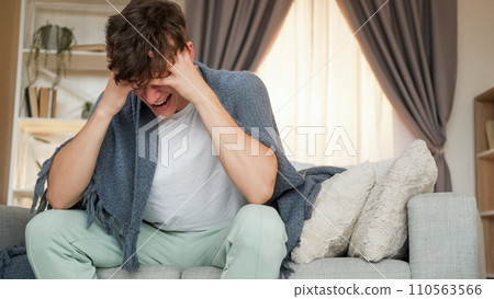 Headache attack. Migraine symptoms. Ill man suffering from unbearable pain grasping head wrapped in blanket sitting on couch at home copy space. Headache attack. Migraine symptoms. Ill man suffering from unbearable pain grasping head wrapped in blanket sitting on couch at home copy space. 110563566
