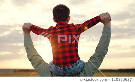 Father carries son on shoulders standing in middle of field at sunset Father carries son on shoulders standing in middle of field at sunset 110563627