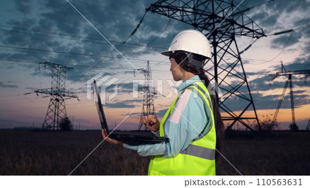 Employee with helmet looks at power transmission lines and types on laptop Employee with helmet looks at power transmission lines and types on laptop 110563631