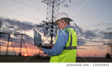 Skilled engineer works on laptop against power transmission lines at sunset Skilled engineer works on laptop against power transmission lines at sunset 110563632