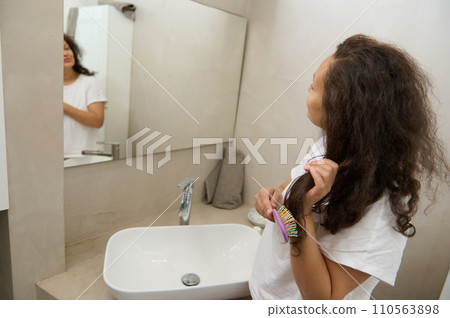 Young woman with long curly brown hair, combing hair with hairbrush, smiling looking at her reflection in the mirror Young woman with long curly brown hair, combing hair with hairbrush, smiling looking at her reflection in the mirror 110563898