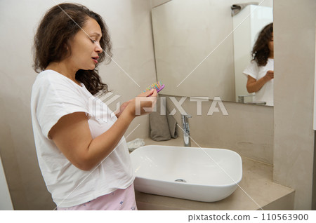 Upset woman removing loss hair from hairbrush after combing her damaged curly and dry hair, standing at sink in bathroom Upset woman removing loss hair from hairbrush after combing her damaged curly and dry hair, standing at sink in bathroom 110563900