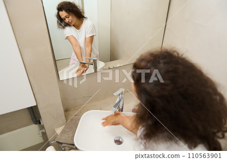 Beautiful woman reflected in the mirror, washing hands with antibacterial soap foam under running water Beautiful woman reflected in the mirror, washing hands with antibacterial soap foam under running water 110563901