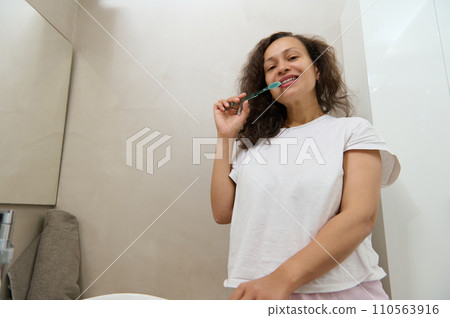Smiling young woman with healthy teeth holding tooth brush, brushing her teeth in bathroom. Dental care and oral hygiene Smiling young woman with healthy teeth holding tooth brush, brushing her teeth in bathroom. Dental care and oral hygiene 110563916
