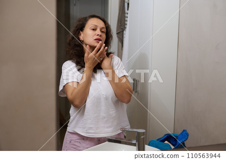 Attractive Latin American young woman applies moisturizer on face and balm on her lips, looking at her mirror reflection 110563944