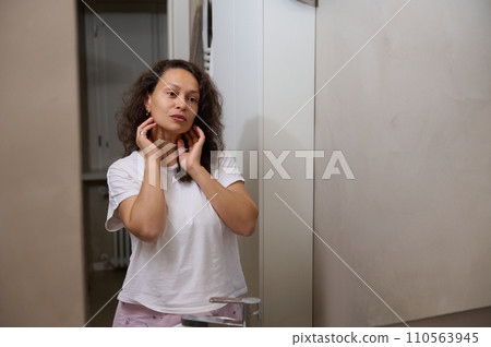 Beautiful Caucasian young woman looking at her mirror reflection in the home bathroom, dressed in pajama 110563945