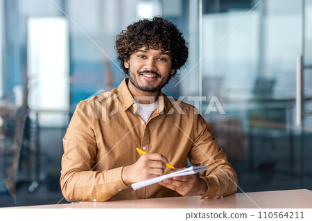 Smiling young male professional with curly hair taking notes at his desk in a contemporary office setting. Smiling young male professional with curly hair taking notes at his desk in a contemporary office setting. 110564211