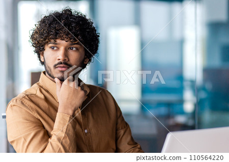 Pensive young businessman with curly hair looks away thoughtfully, portraying determination and strategy planning in a contemporary workspace. 110564220