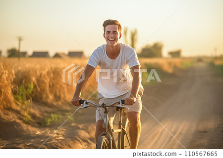 A young European man rides a bicycle along a country road past a wheat field, smiles happily and greets the dawn 110567665