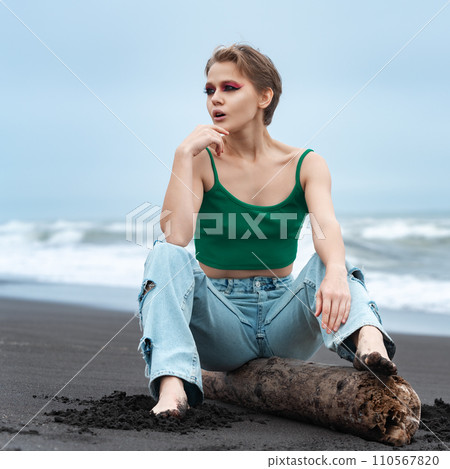Blonde woman with bright makeup is sitting on log on beach, looking away with hand resting on chin Blonde woman with bright makeup is sitting on log on beach, looking away with hand resting on chin 110567820
