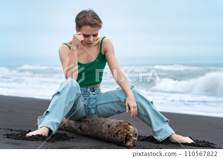 Woman sitting on log with legs spread and head lowered, looking down on beach of Pacific Ocean Woman sitting on log with legs spread and head lowered, looking down on beach of Pacific Ocean 110567822