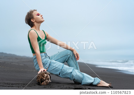 Awesome hipster woman with eyes closed leaning back on log on black sandy beach of Pacific Ocean 110567823
