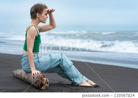 Hipster woman sitting on log on black sandy beach of ocean, raised hand to forehead, looking at view 110567824