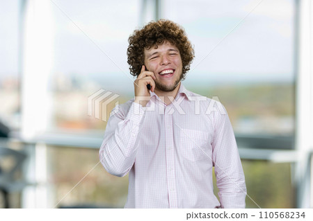 Portrait of a young man in white shirt talking on the phone and laughing. Indoor window in the background. 110568234