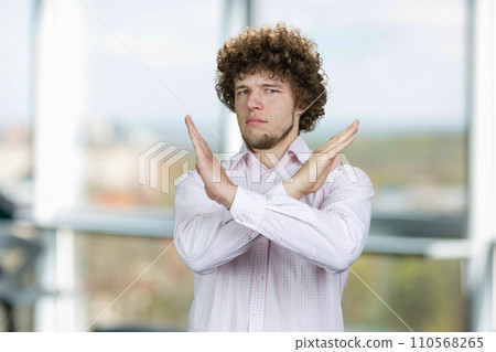 Portrait of a young man with curly hair cross his arms as sign of prohibition. Office window in the background. 110568265