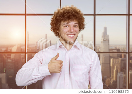 Portrait of a happy young man with curly hair shows thumb up. Checkered window with cityscape view. Portrait of a happy young man with curly hair shows thumb up. Checkered window with cityscape view. 110568275