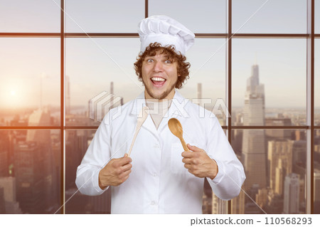 Portrait of a happy young caucasian male cook with a wooden spoon. Checkered window with cityscape in the background. Portrait of a happy young caucasian male cook with a wooden spoon. Checkered window with cityscape in the background. 110568293