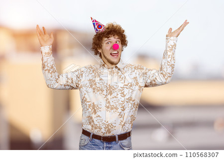 Happy young guy with clown nose fooling around making funny faces. Blurred cityscape in the background. 110568307