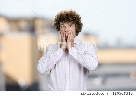 Portrait of shocked amazed young guy with curly hair touching his face. Blurred urban background. Portrait of shocked amazed young guy with curly hair touching his face. Blurred urban background. 110568316