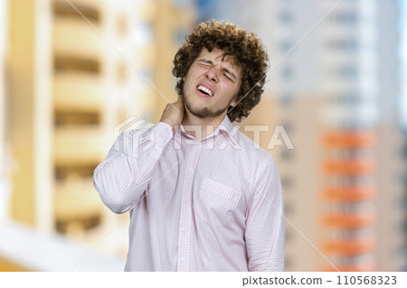 Young handsome man with curly hair having neckache. Residential area in the background. Young handsome man with curly hair having neckache. Residential area in the background. 110568323