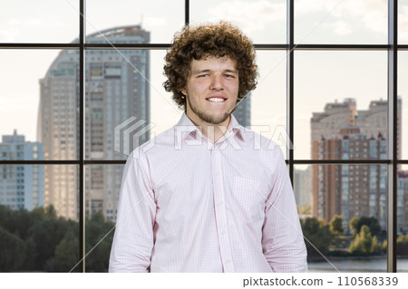 Happy young attractive man with curly hair bites his lip. Indoor window in the background. 110568339