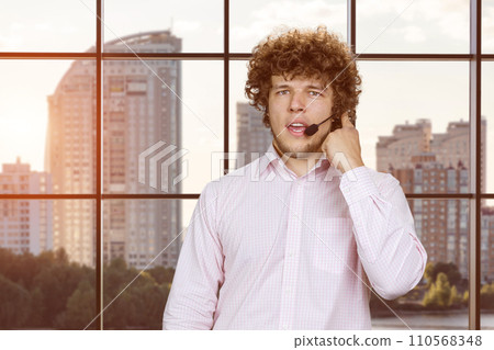 Portrait of a young serious confident successful man talking in microphone. Indoor window with evening cityscape view in the background. 110568348