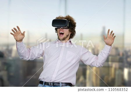 Portrait of young excited man with curly hair experiencing virtual reality. Blurred cityscape in the background. 110568352