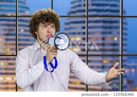 Handsome young man with curly hair in white shirt speaks in megaphone. Indoor window with night city view in the background. 110568366