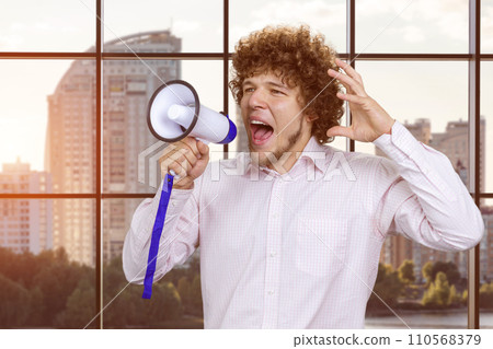 Portrait of a young excited expressive caucasian man with curly hair in white shirt screaming hot news shout in megaphone. Indoor window in the background. 110568379