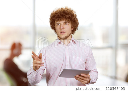 Portrait of a young businessman with curly hair standing with tablet pc device pointing at something. Office environment in the background. Portrait of a young businessman with curly hair standing with tablet pc device pointing at something. Office environment in the background. 110568403