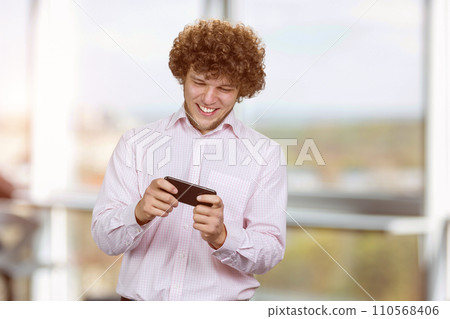 Excited happy young caucasian male gamer playing using smartphone. Guy playing mobile videogame app or watching digital video stream holding cell phone indoors. Excited happy young caucasian male gamer playing using smartphone. Guy playing mobile videogame app or watching digital video stream holding cell phone indoors. 110568406