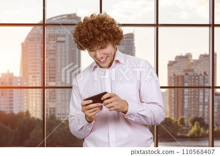 Excited happy young caucasian man gamer playing using smartphone. Guy playing mobile videogame app or watching a video stream holding cell phone indoors. Excited happy young caucasian man gamer playing using smartphone. Guy playing mobile videogame app or watching a video stream holding cell phone indoors. 110568407