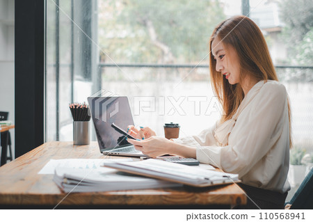 Attractive young woman working on the mobile phone and smiling while sitting at her working place in office and looking at laptop 110568941